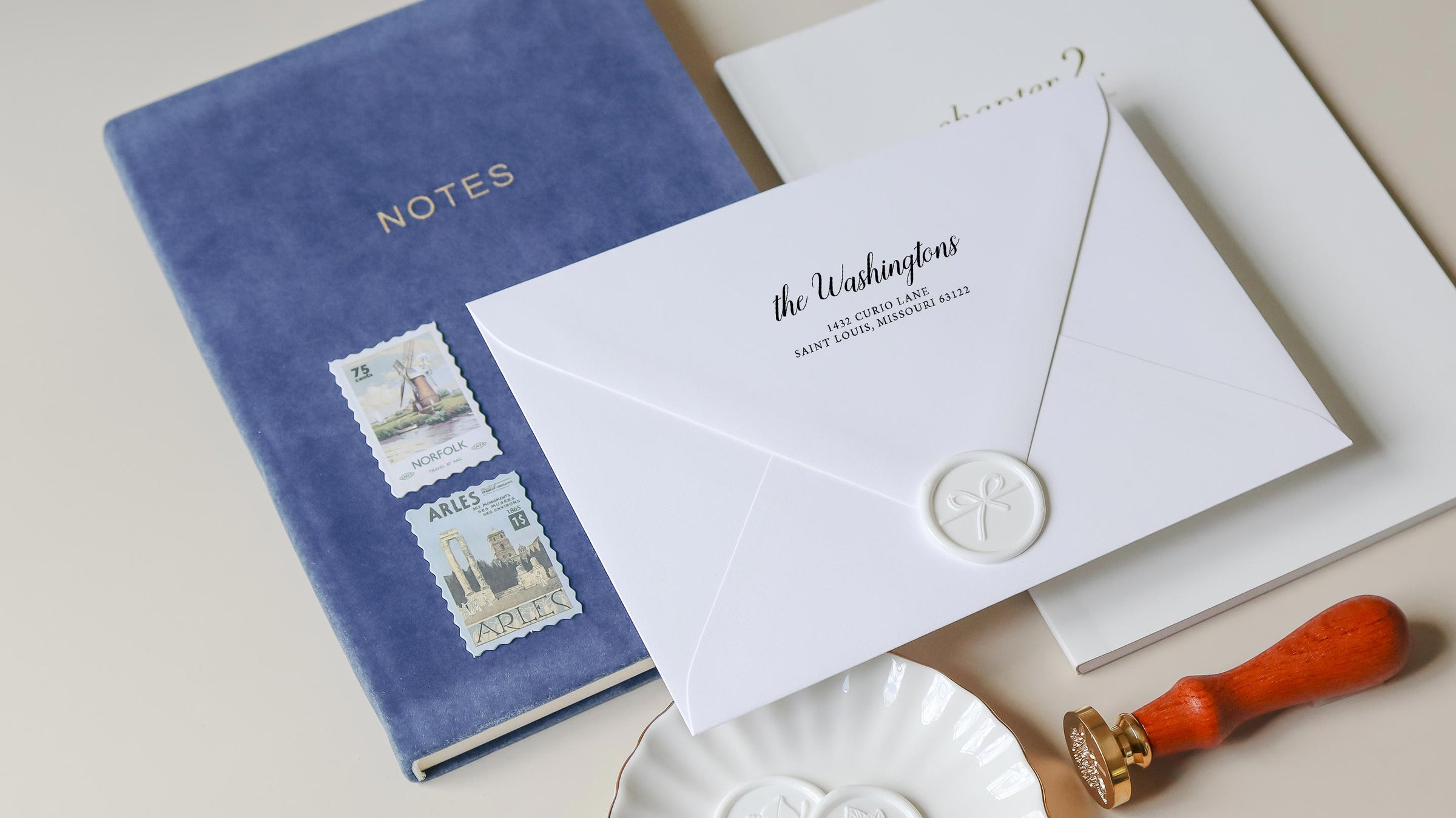 A white envelope with custom address stamp sits on a blue velvet notebook with vintage stamps and a wax seal