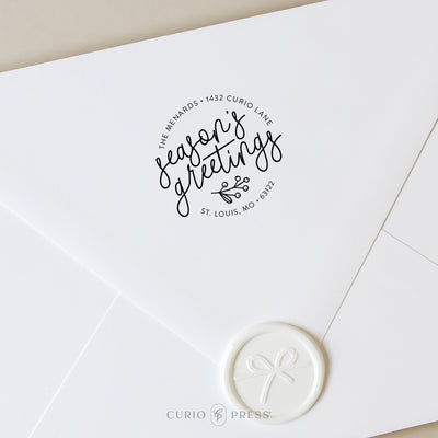 Custom holiday stamp with "Season's Greetings" in a whimsical script paired with a holly sprig stamped on a white envelope
