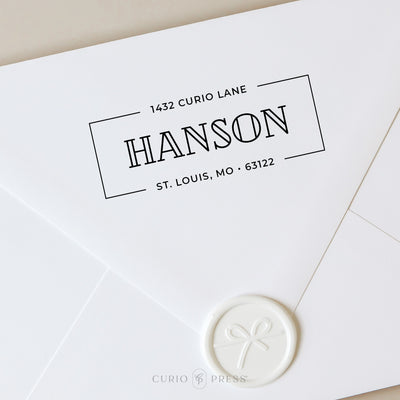 Custom address stamp with bold outlined last name in center and street address and city details in top and bottom of the frame