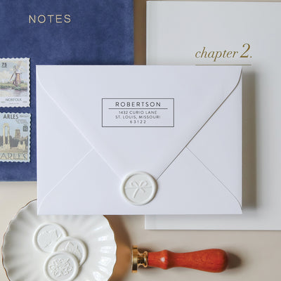 White envelope with modern address stamp and wax seal, surrounded by stationery items on a neutral background.