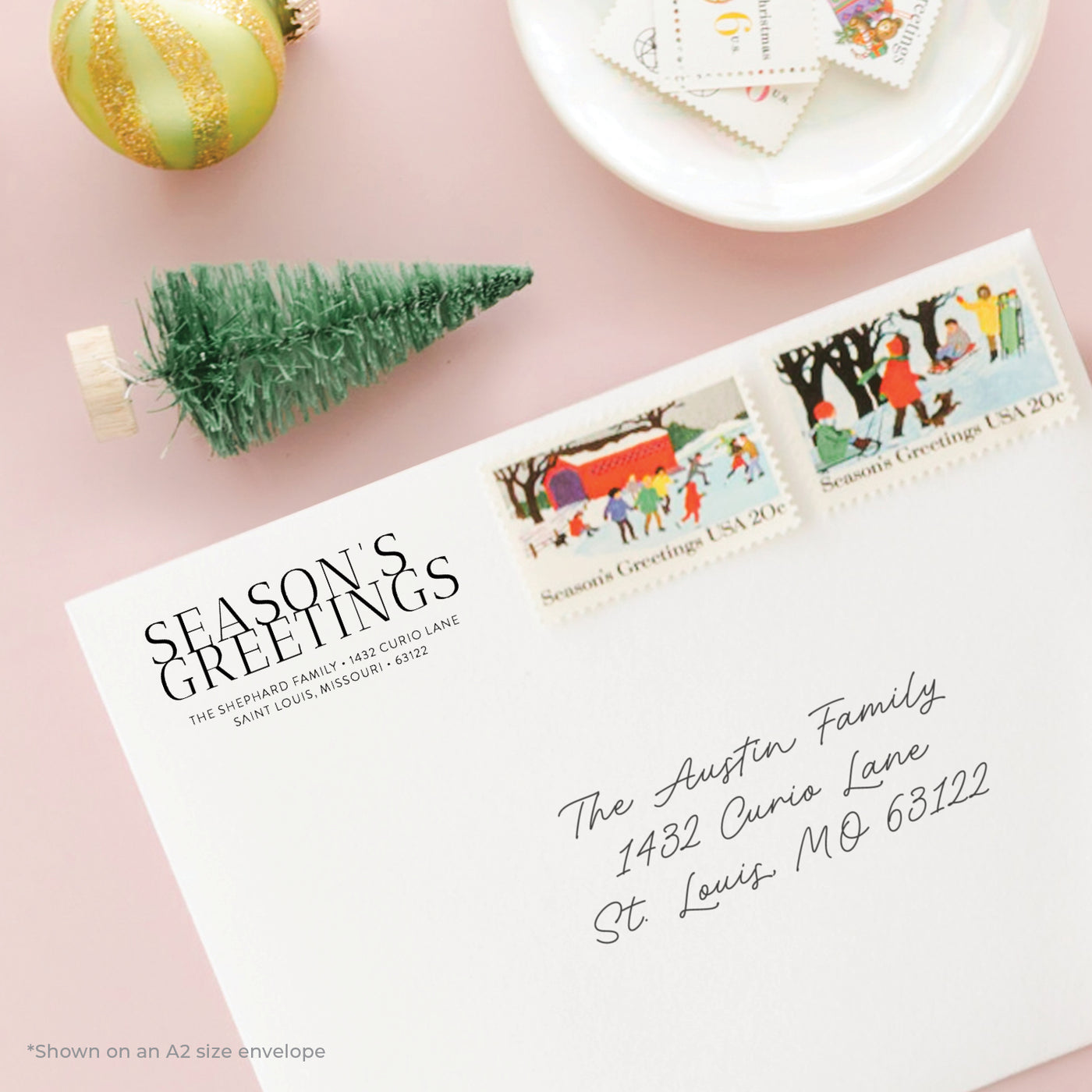Holiday-themed personalized address stamp on envelope with Season's Greetings stamps on a pink background